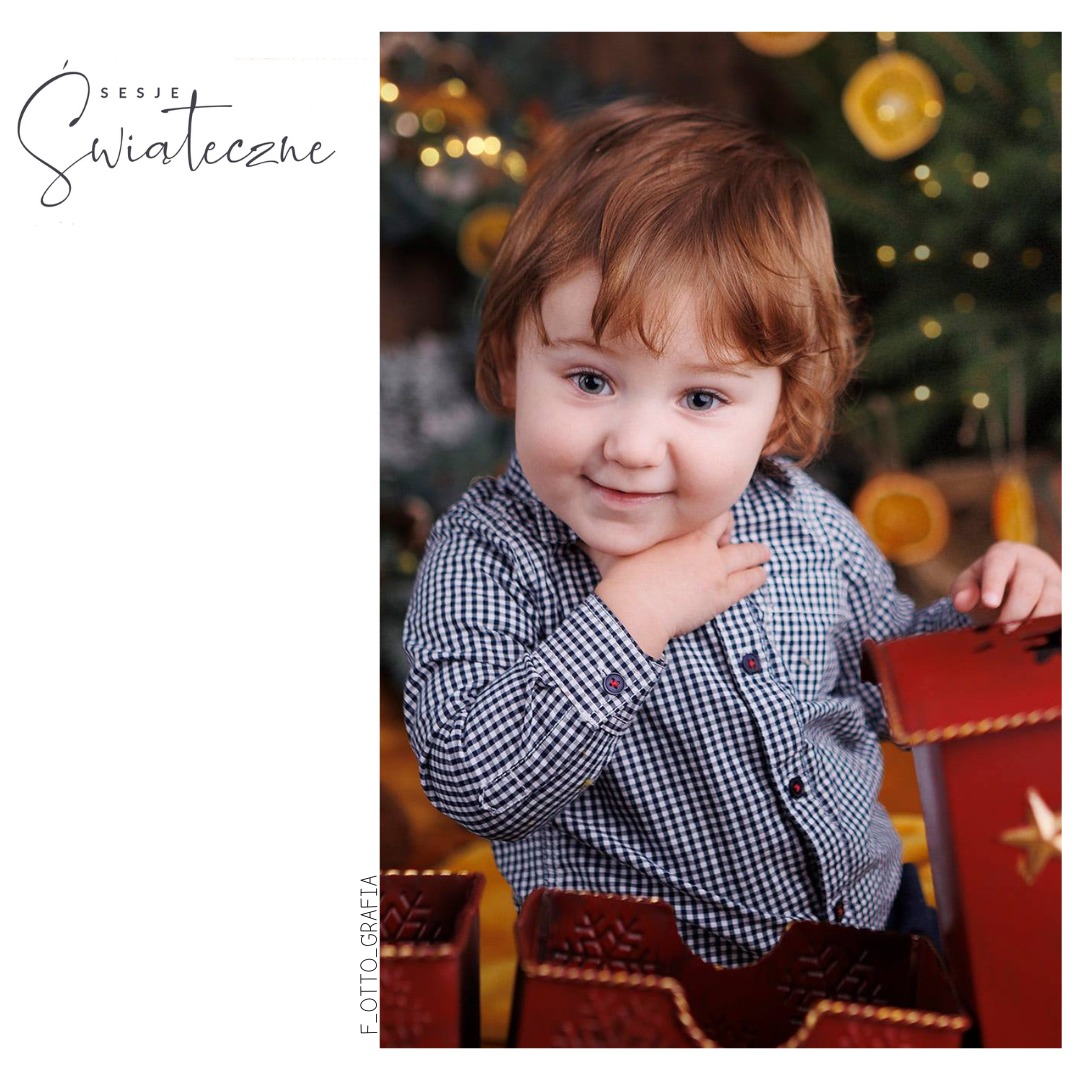 Uśmiechnięty chłopiec w koszuli w kratę pozuje na tle choinki z dekoracjami i drewnianych sanek. Sesja świąteczna, ciepłe oświetlenie.