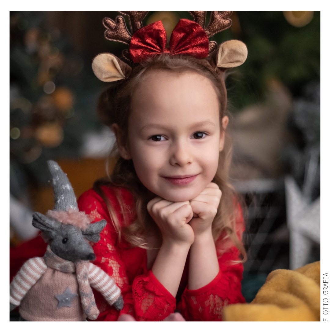 Portret uśmiechniętej dziewczynki w stroju renifera z pluszową zabawką na tle świątecznych dekoracji, w ciepłej tonacji kolorystycznej.