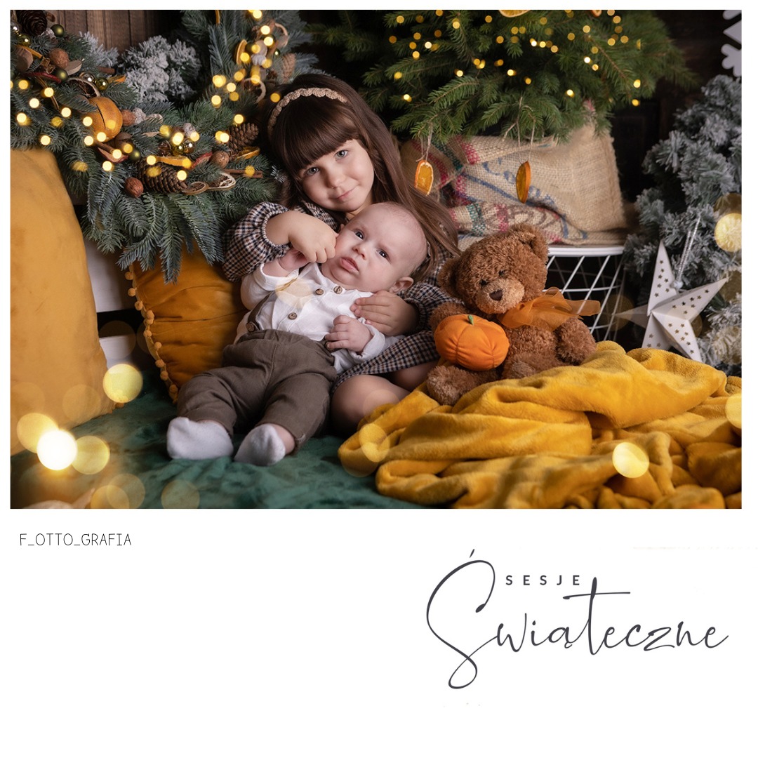 Uśmiechnięta dziewczynka obejmuje niemowlę na świątecznej sesji zdjęciowej z pluszowym misiem i dekoracjami bożonarodzeniowymi w ciepłych, naturalnych barwach.