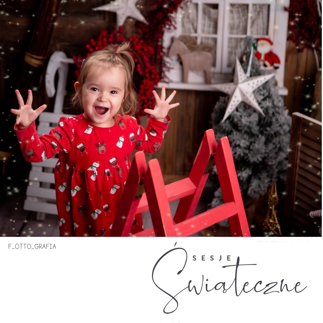 Radosna dziewczynka w czerwonej sukience z motywem świątecznym, pozująca na tle bożonarodzeniowej scenografii z sanek i choinki.