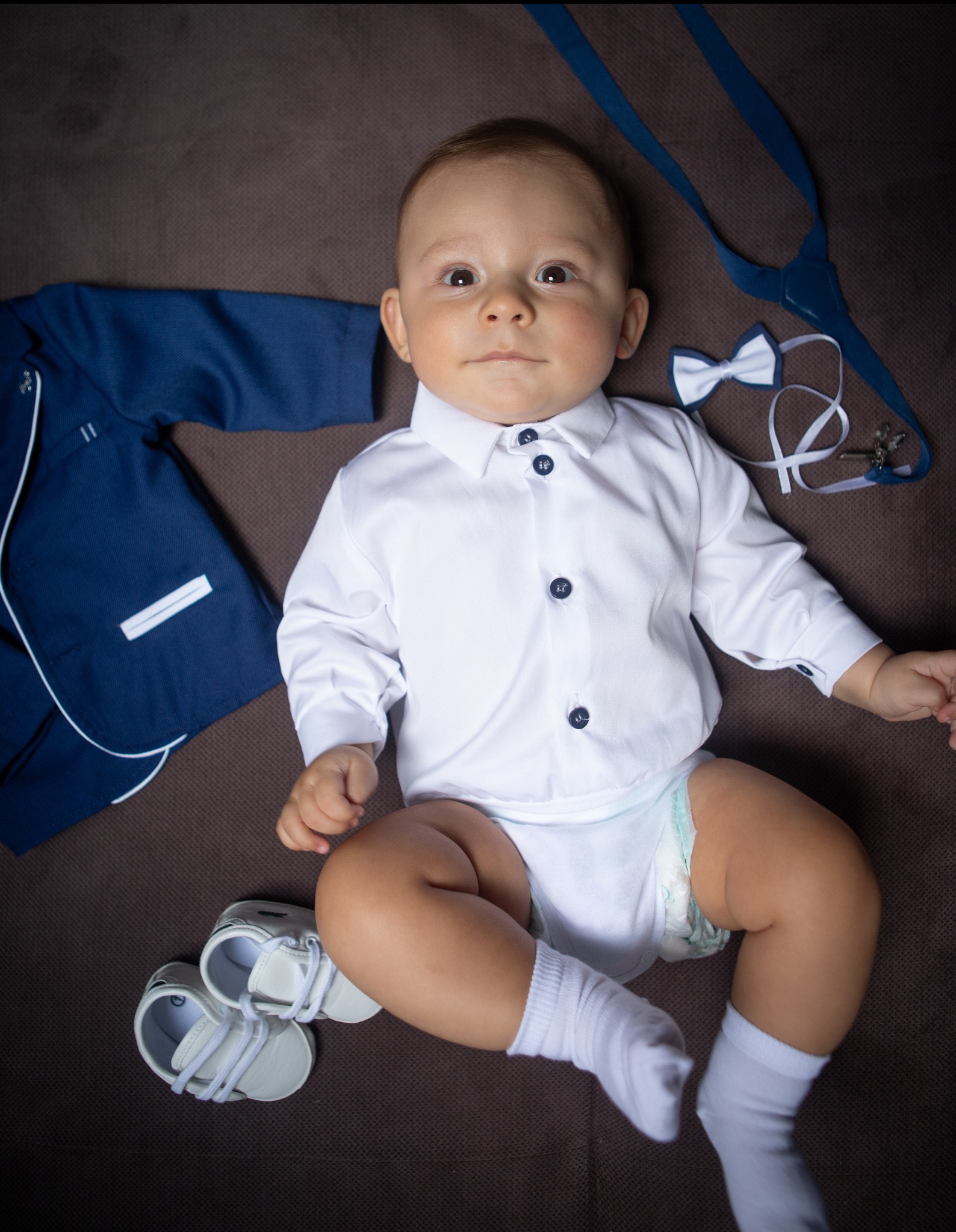 Uroczy niemowlak w białej koszuli z granatowymi guzikami, leżący obok miniaturowego garnituru, muchy i bucików na brązowym tle.