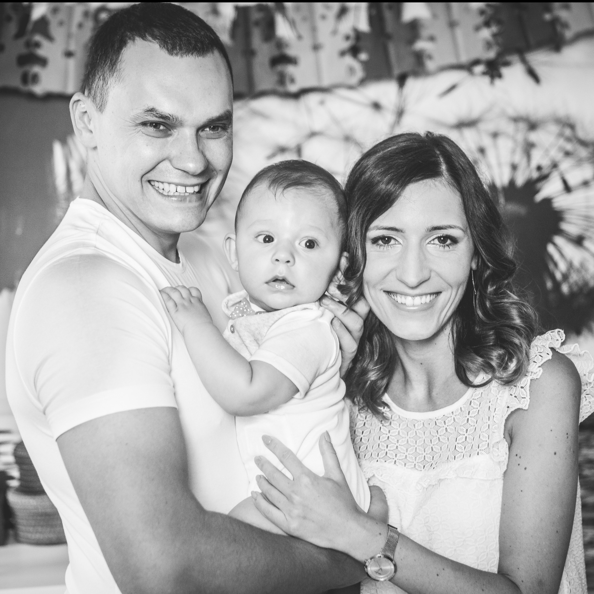 Czarno-białe ujęcie uśmiechniętej rodziny: tata trzyma niemowlę, obok mama w koronkowej sukience, w tle abstrakcyjna dekoracja.