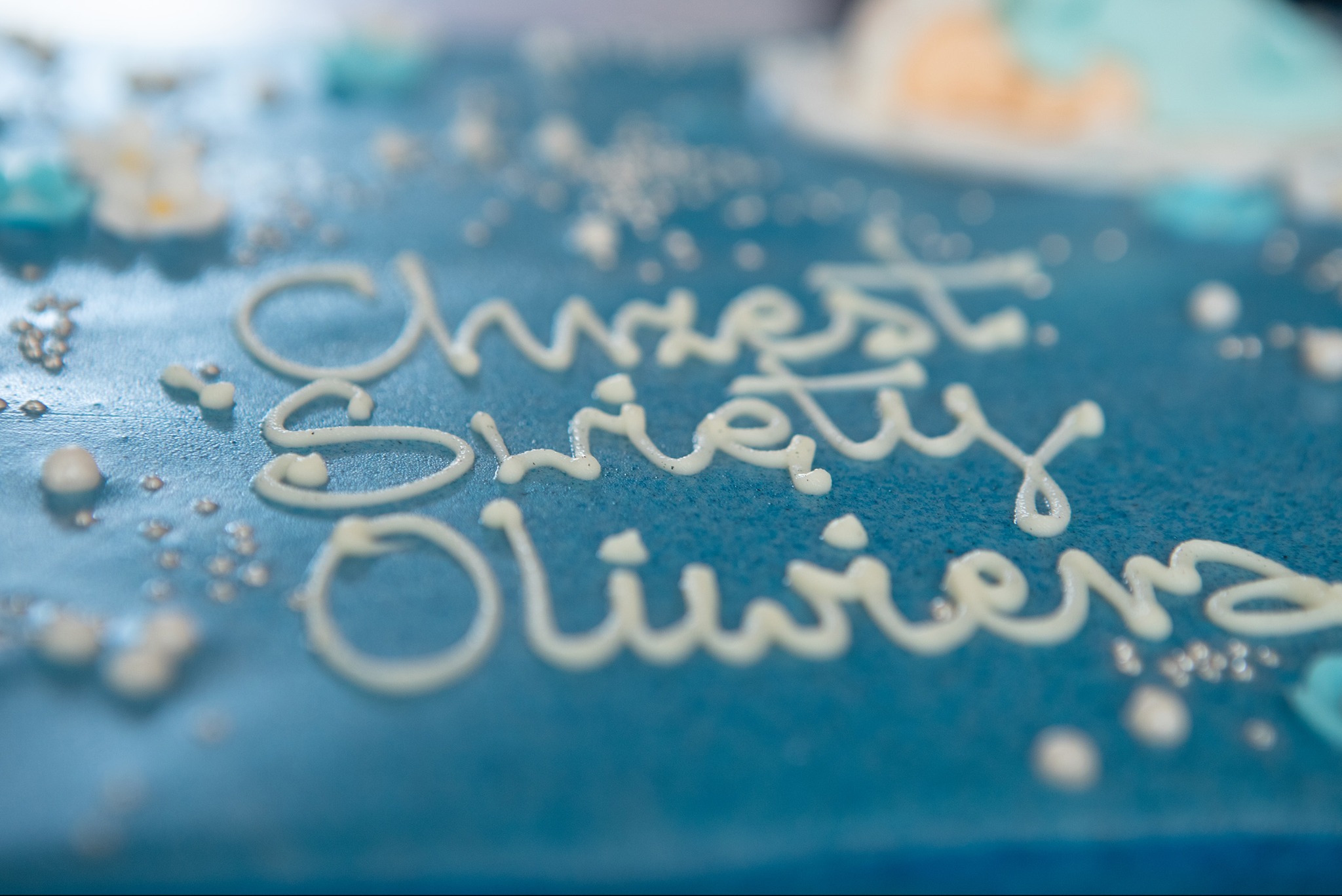 Błękitny tort z białym napisem 'Chrzest Święty Oliwiera', zdobiony perłowymi koralikami i kwiatkami. Ujęcie z bliska, delikatne rozmycie tła.