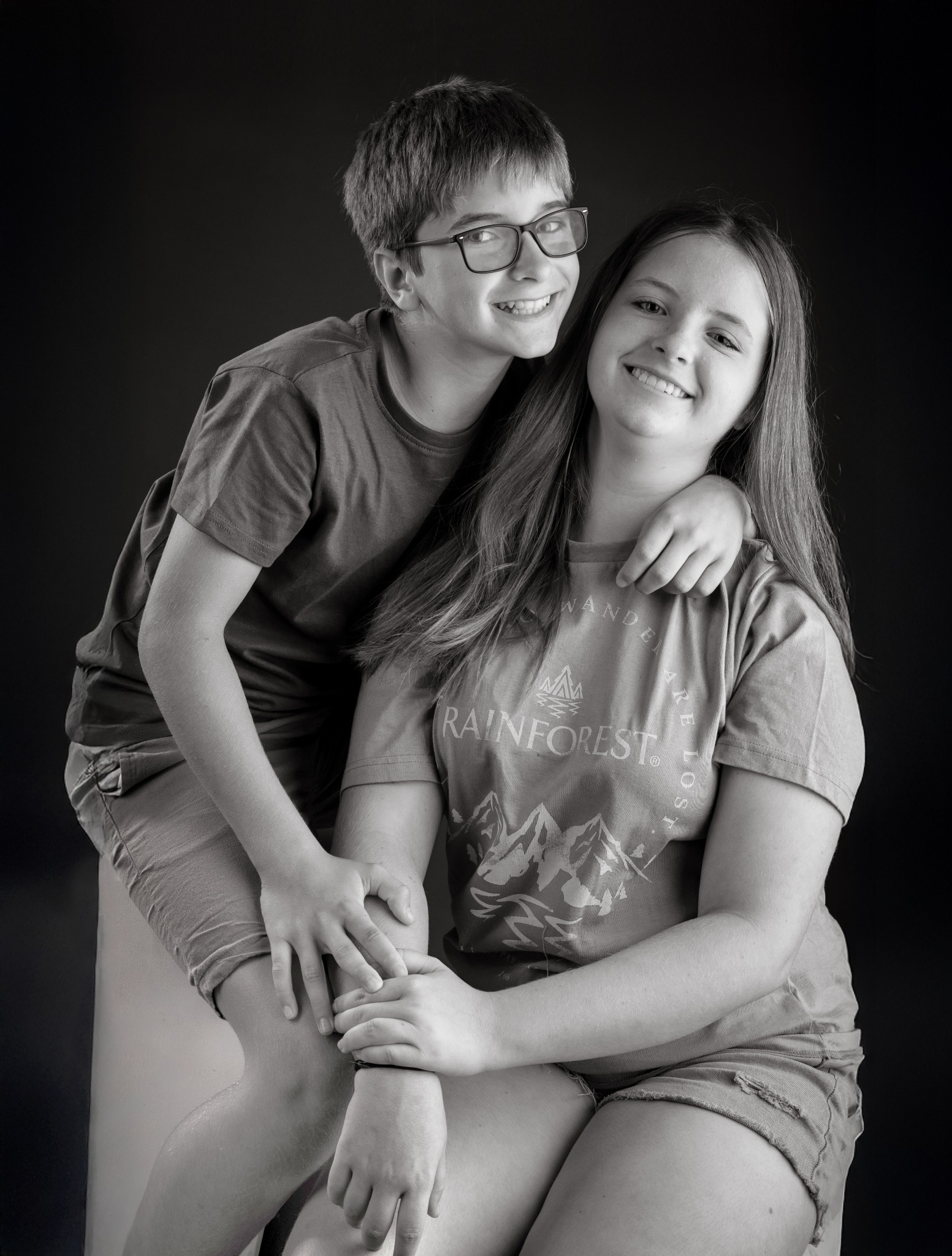 Czarno-białe portretowe ujęcie uśmiechniętego rodzeństwa: nastolatki siedzącej z chłopcem opierającym się o jej ramię, na ciemnym tle.