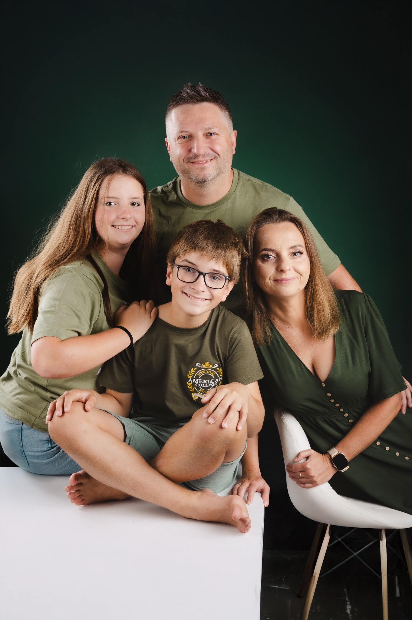 Portret rodzinny: uśmiechnięci rodzice z dwójką dzieci w zielonych ubraniach pozują na białym stole na tle ciemnozielonego tła w studio fotograficznym.