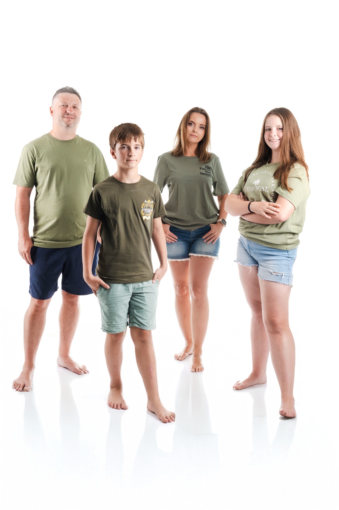 Rodzinna sesja na białym tle: ojciec w zielonej koszulce, syn z rękami w kieszeniach, mama i córka w dżinsowych szortach. Uśmiechnięci, boso.