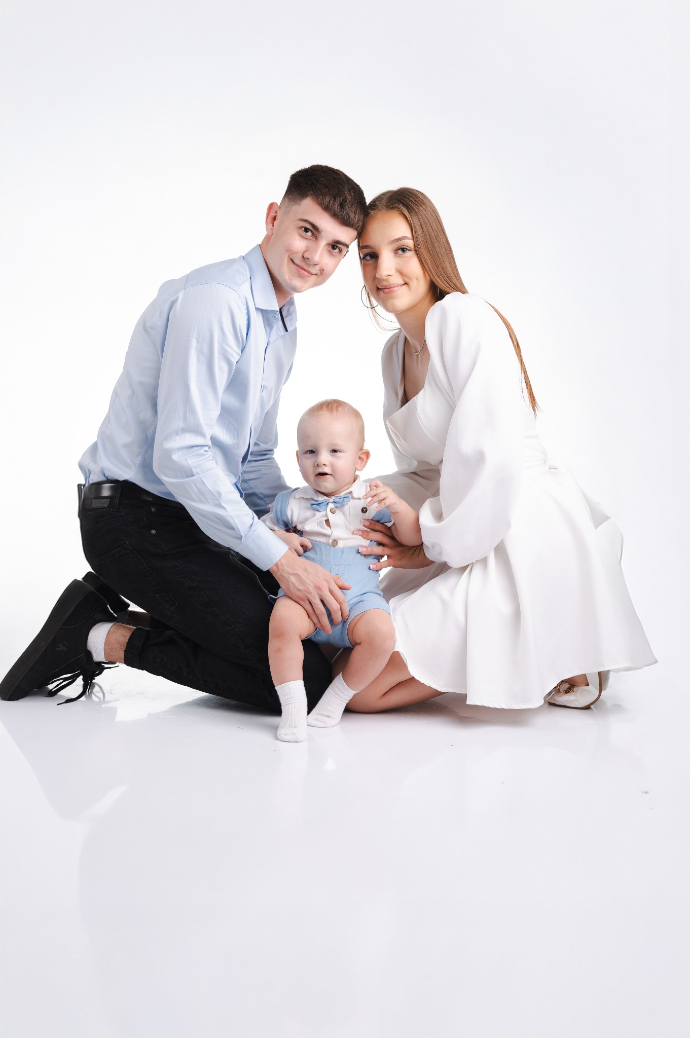 Uśmiechnięta rodzina: tata na kolanach w błękitnej koszuli, mama w białej sukience i synek w błękitnym ubranku, pozują na białym tle w studio.