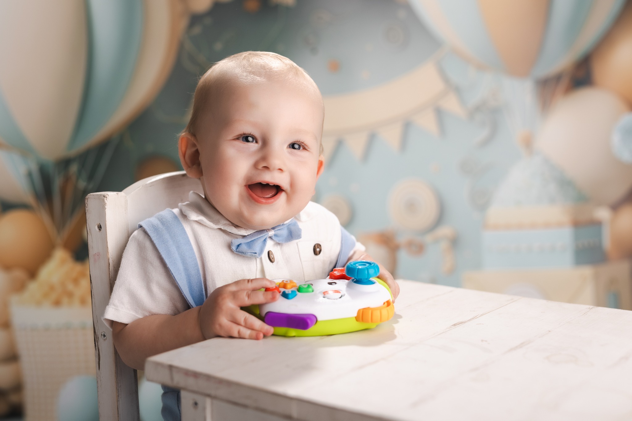 Uśmiechnięty niemowlak w błękitnym ubranku siedzi przy stole, trzymając zabawkę na tle pastelowej dekoracji z motywem balonów.