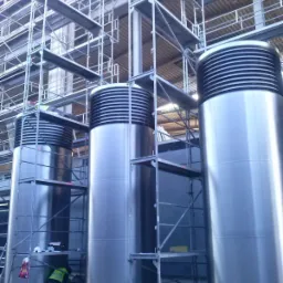 Trzy duże, srebrne, cylindryczne kanały wentylacyjne z poziomymi żebrami u góry, ustawione pionowo wewnątrz budynku w trakcie montażu, otoczone rusztowaniami i pracownikami w kamizelkach odblaskowych.