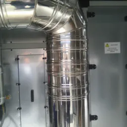 Błyszczące, metalowe rury wentylacyjne połączone pod kątem prostym, z widocznym fragmentem urządzenia wentylacyjnego i ostrzeżeniem w języku niemieckim.