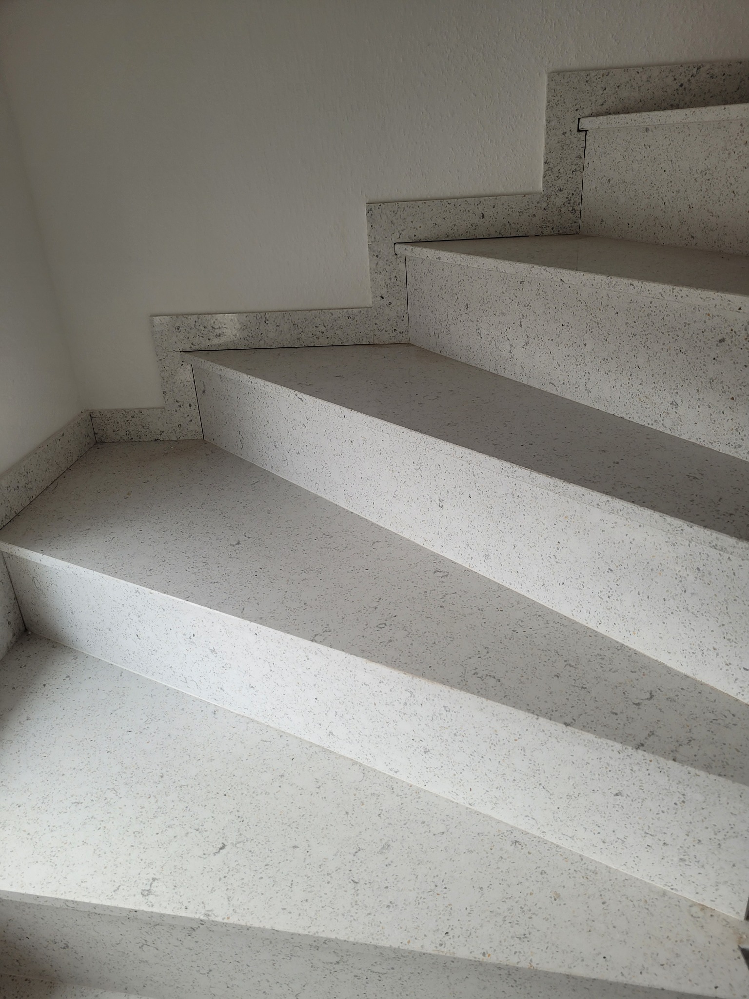 Schody wewnętrzne wykończone jasnym, nakrapianym kamieniem kompozytowym, z widocznymi detalami obróbki krawędzi i minimalistycznym połączeniem ze ścianą.