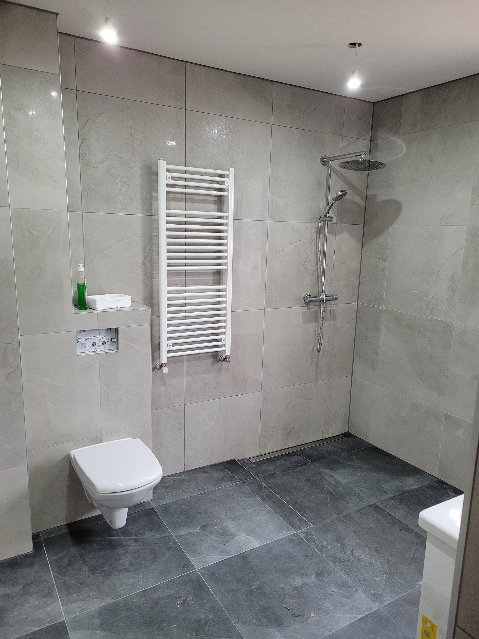 Nowoczesna łazienka z szarymi płytkami imitującymi kamień na ścianach i podłodze, podwieszana toaleta, biały grzejnik drabinkowy i kabina prysznicowa typu walk-in z deszczownicą.