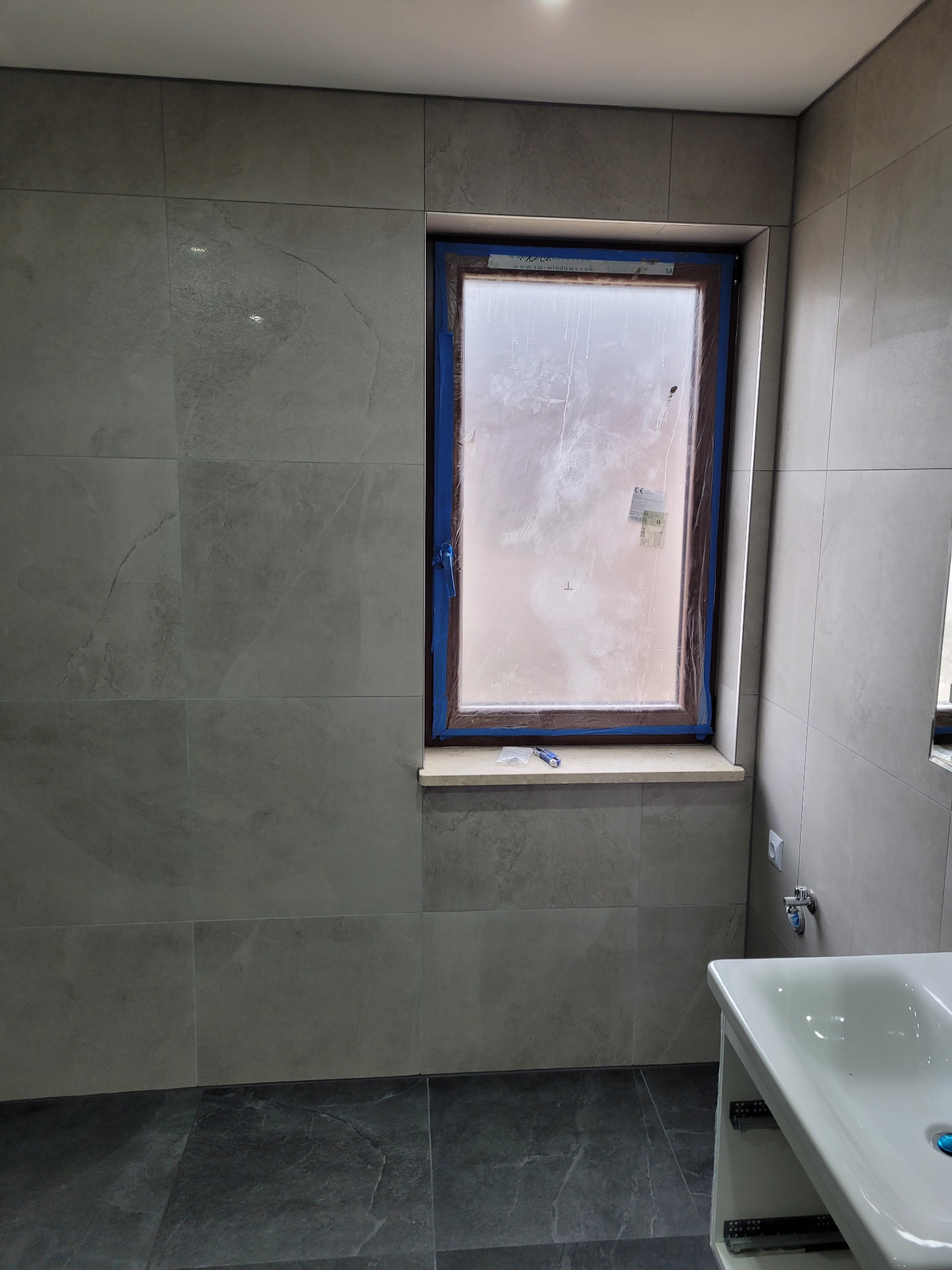 Łazienka w trakcie wykańczania: szare płytki ścienne i podłogowe, okno z zabezpieczającą folią i niebieską taśmą malarską, biała umywalka z szafką.