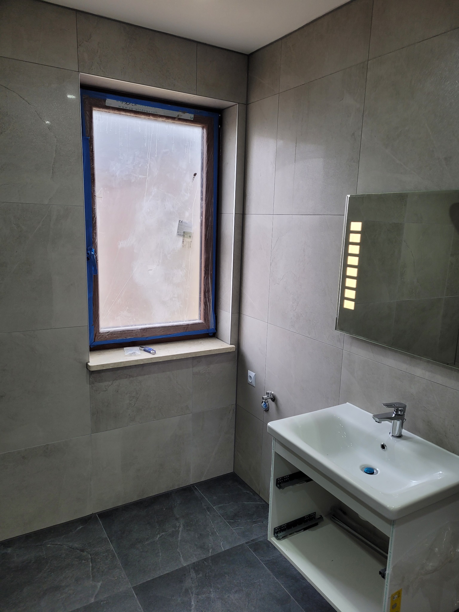 Nowoczesna łazienka w trakcie wykańczania: szare płytki na ścianach i podłodze, biała umywalka z szafką, lustro z podświetleniem LED, okno zabezpieczone folią malarską, widoczne narzędzia...