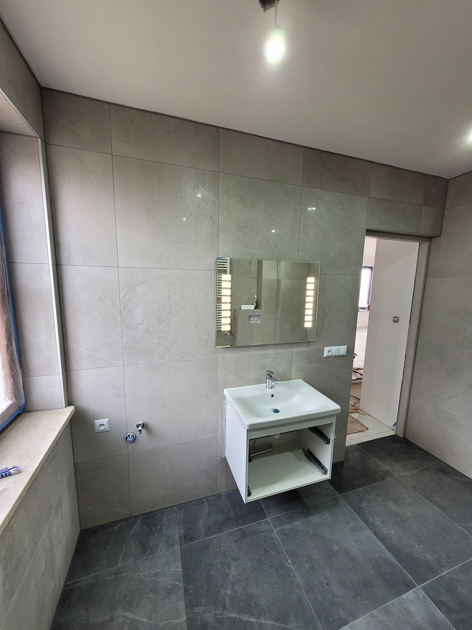 Wykończona łazienka z szarymi płytkami imitującymi kamień na ścianach i podłodze, podwieszana biała szafka z umywalką i lustro z oświetleniem LED. Widoczne gniazdka elektryczne i rura...