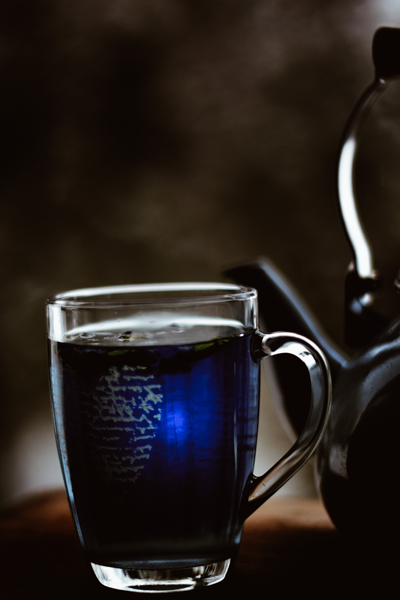 Szklanka z niebieskim napojem i ziołami, obok czarny czajnik na tle ciemnego, rozmytym tle.