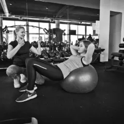 Trener personalny nadzoruje ćwiczenia brzuszków na piłce fitness w siłowni, instruując gestami. W tle widoczne ciężary i lustra.