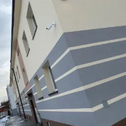 Odnowiona elewacja budynku wielorodzinnego z poziomymi pasami w odcieniach szarości i beżu, widoczna kamera monitoringu, okna z jasnymi ramami i fragment chodnika z resztkami śniegu.