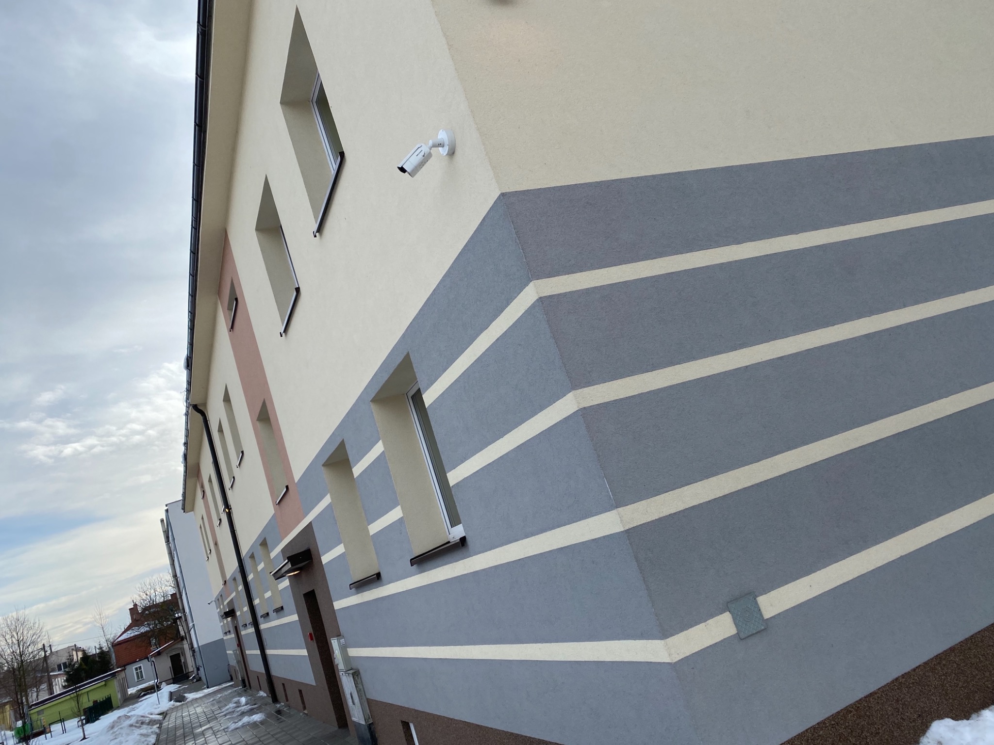 Odnowiona elewacja budynku wielorodzinnego z poziomymi pasami w odcieniach szarości i beżu, widoczna kamera monitoringu, okna z jasnymi ramami i fragment chodnika z resztkami śniegu.