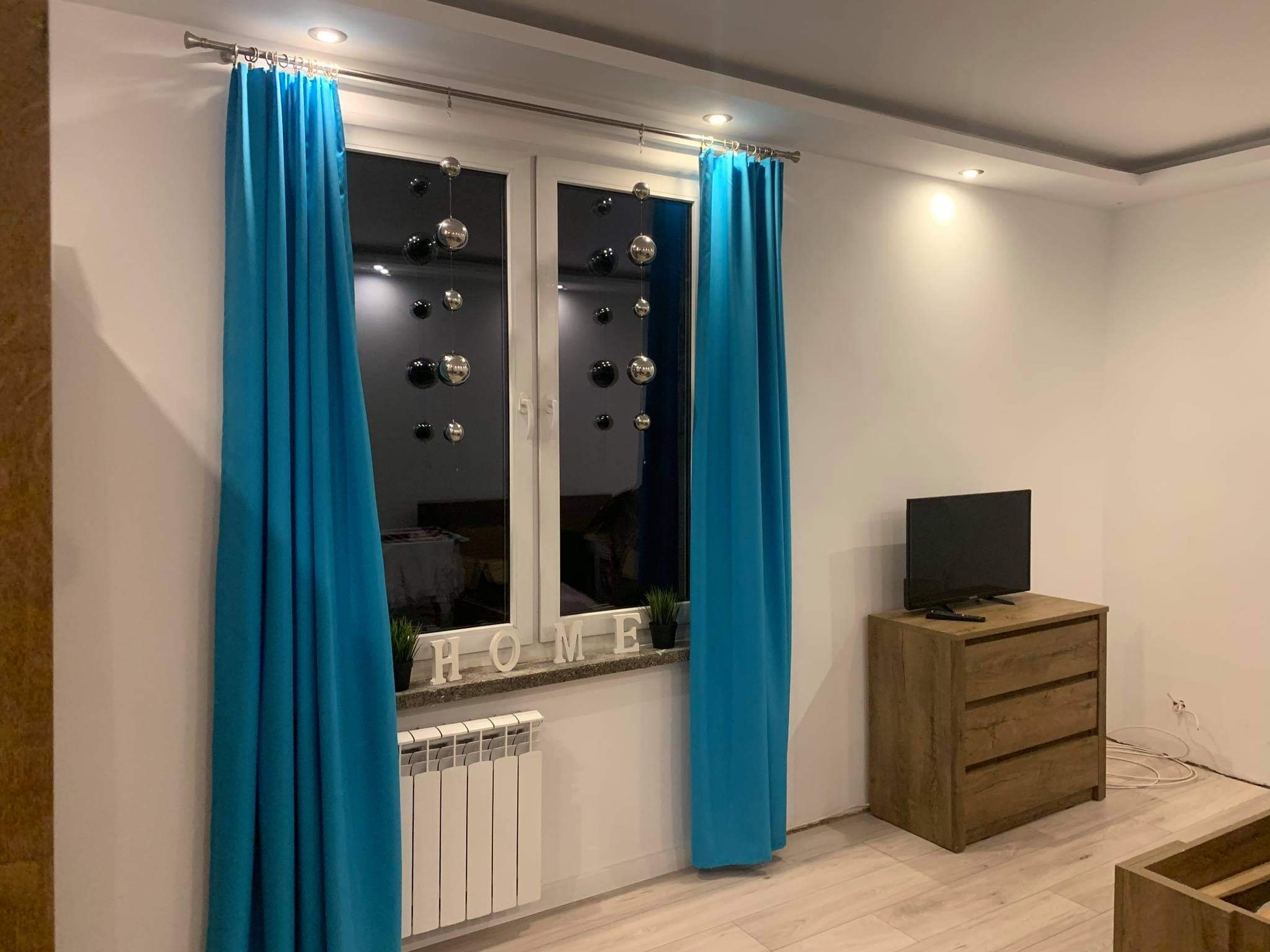 Pokój po odświeżeniu: turkusowe zasłony na karniszu, okno z dekoracjami w postaci srebrnych i czarnych bombek, biały grzejnik, drewniana komoda z telewizorem, podłoga wyłożona panelami.