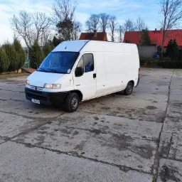 Biały, dostawczy bus marki Peugeot zaparkowany na betonowym placu w słoneczny dzień. Widoczne drzewa i budynki w tle.