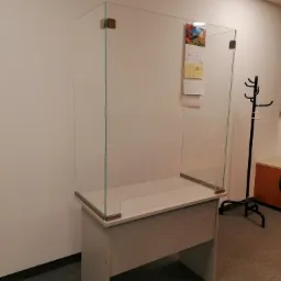 Przezroczysta szklana osłona na biurko z metalowymi mocowaniami, chroniąca przed kontaktem, z kalendarzem przyczepionym do szyby, w tle wieszak na ubrania i jasne ściany.