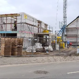 Dwa budynki w stanie surowym otwartym, jeden z widocznym szkieletem dachu, oba otoczone rusztowaniami i siatką zabezpieczającą teren budowy, z paletami i materiałami budowlanymi w pobliżu.