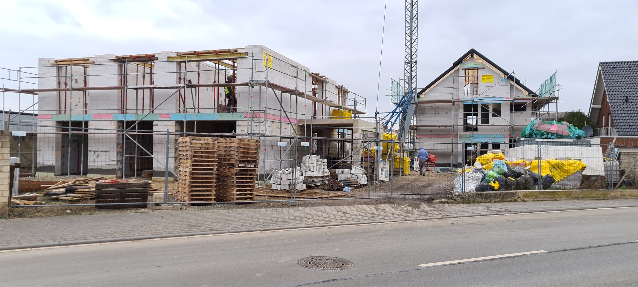 Dwa budynki w stanie surowym otwartym, jeden z widocznym szkieletem dachu, oba otoczone rusztowaniami i siatką zabezpieczającą teren budowy, z paletami i materiałami budowlanymi w pobliżu.