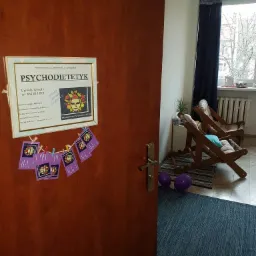 Tabliczka na drzwiach Pracowni Psychodietetyki Urszuli Wesoły z powieszonymi wizytówkami, widoczny fragment poczekalni z dwoma drewnianymi leżakami i fioletowymi balonami.