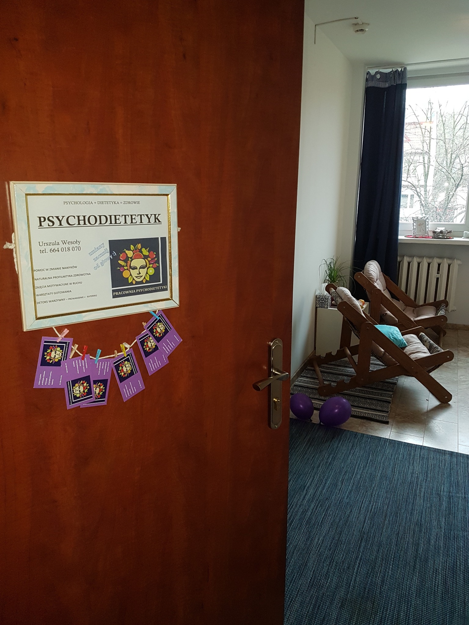 Tabliczka na drzwiach Pracowni Psychodietetyki Urszuli Wesoły z powieszonymi wizytówkami, widoczny fragment poczekalni z dwoma drewnianymi leżakami i fioletowymi balonami.