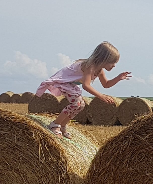 Dziewczynka w różowej sukience i legginsach w truskawki skacze po belach siana na polu w słoneczny dzień.