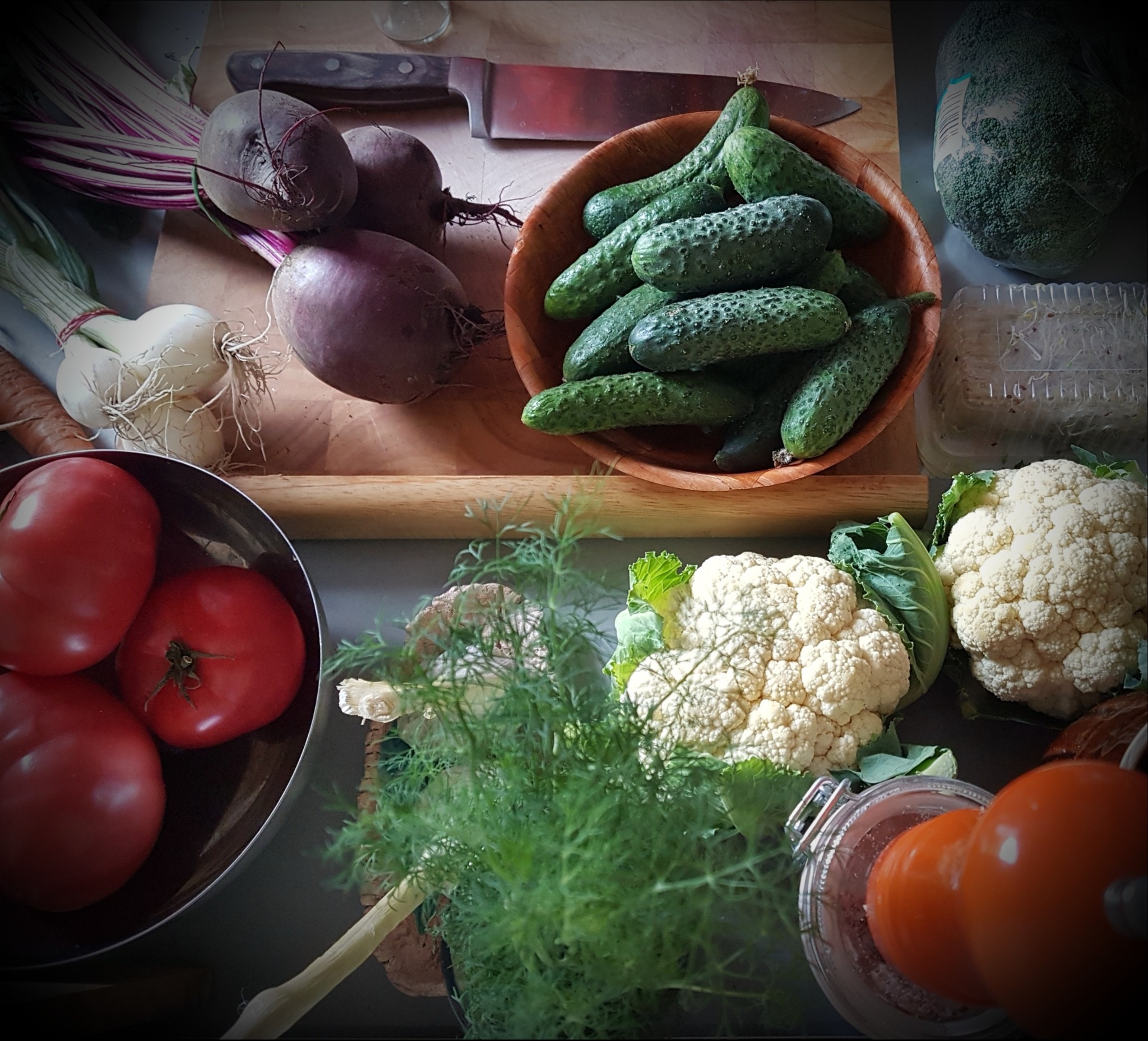 Kompozycja świeżych warzyw: buraki z liśćmi, cebula, czosnek, pomidory w metalowej misce, ogórki w drewnianej miseczce, brokuły w folii, kiełki, dwa kalafiory, pęczek koperku, nóż i słoik.