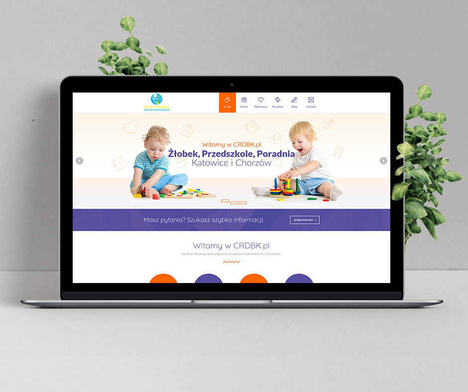 Ekran laptopa prezentujący stronę internetową żłobka i przedszkola CRDBK.pl z Katowic i Chorzowa, z dziećmi bawiącymi się na sliderze i zieloną rośliną po obu stronach urządzenia.