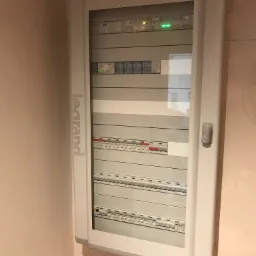 Zamknięta skrzynka elektryczna marki Legrand, zamontowana w rogu pomieszczenia, z widocznymi bezpiecznikami i włącznikami oraz zielonymi diodami sygnalizacyjnymi.