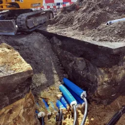 Żółta koparka gąsienicowa JCB przy wykopie z ułożonymi niebieskimi rurami kablowymi w osłonie, widoczne kable i uszczelnienia, w tle hałda ziemi i budynki.