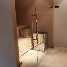 Szklane drzwi do nowej sauny z drewnianymi ławkami i piecem Harvia, widoczne na tle marmurowej podłogi.