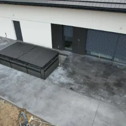 Zewnętrzna powierzchnia z szarego betonu architektonicznego okalająca wbudowane jacuzzi z czarną pokrywą, widok z góry na tle nowoczesnego domu z białymi ścianami i szarymi roletami.