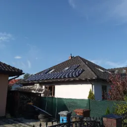 Dom jednorodzinny z panelami słonecznymi na dachu, widok zza ogrodzenia, błękitne niebo z delikatnymi chmurami w tle.