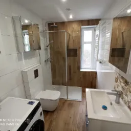 Nowoczesna łazienka z kabiną prysznicową typu walk-in, wiszącą toaletą, umywalką z szafką i pralką, wykończona płytkami imitującymi drewno i białymi płytkami z dekoracyjnym pasem z motywem liści.