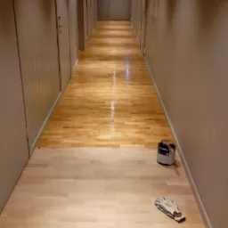 Malowanie podłogi w korytarzu; widoczna pomalowana i niepomalowana część podłogi, puszka farby i rękawice robocze.