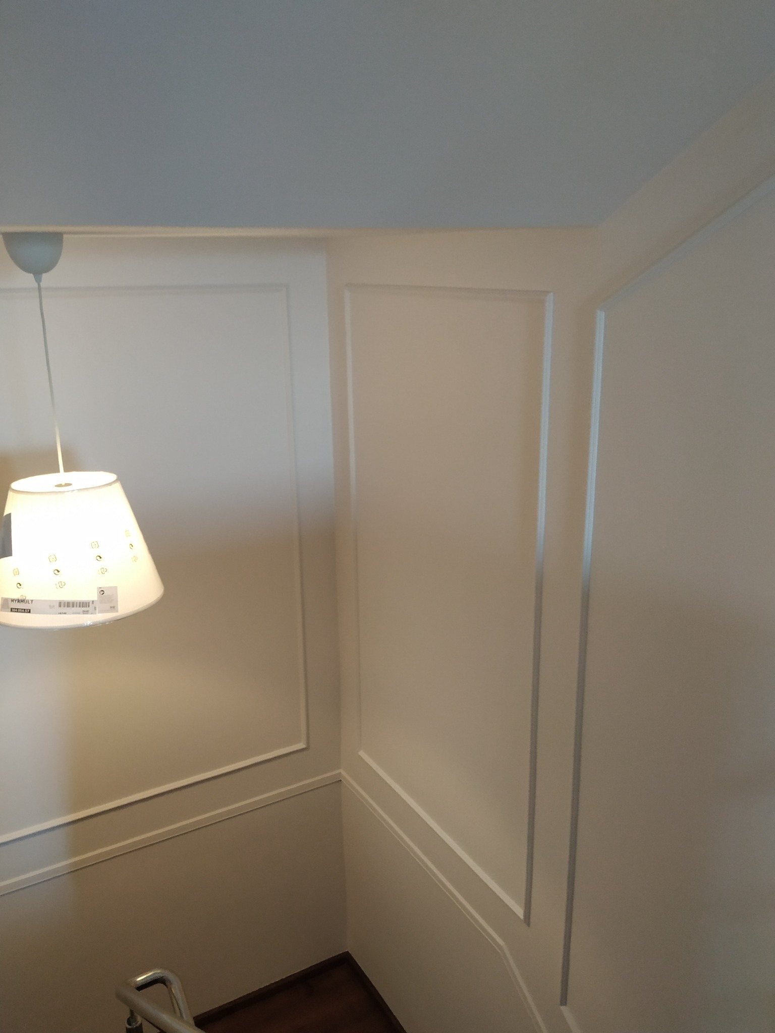Ściana klatki schodowej z dekoracyjnymi listwami i lampą wiszącą IKEA, widoczna część schodów z metalową barierką.