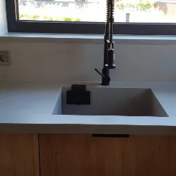 Szary blat kamienny z wbudowanym zlewem i czarną baterią kuchenną na tle okna w minimalistycznej kuchni.