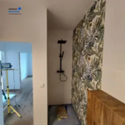 Nowoczesna łazienka po remoncie z czarną armaturą prysznicową i ścianą pokrytą tapetą w motywy roślinne. W rogu widoczny worek z materiałem budowlanym i lampa na statywie.