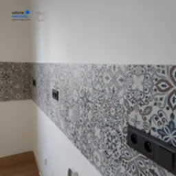 Ściana w łazience wykończona dekoracyjnymi płytkami ceramicznymi w stylu marokańskim, z widocznymi gniazdkami elektrycznymi w kolorze czarnym. Biała ściana powyżej płytek. Ujęcie z boku, ukazujące...