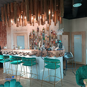 Nowoczesny bar w lokalu usługowym z blatem z naturalnego drewna, podświetlaną ścianą z butelkami, wiszącą instalacją artystyczną z drewnianych elementów i turkusowymi krzesłami barowymi.