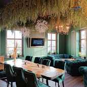 Eleganckie wnętrze restauracji z zielonymi ścianami i sufitem zdobionym roślinami, masywny drewniany stół otoczony krzesłami z zielonego aksamitu, w tle sofy w stylu Chesterfield w tym samym...