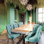 Elegancka jadalnia z długim, drewnianym stołem z surowymi krawędziami, otoczonym turkusowymi krzesłami z pikowanym oparciem. Zielone ściany i metalowe akcenty tworzą nowoczesny, ale przytulny...