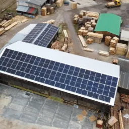 Widok z góry na budynki przemysłowe z panelami fotowoltaicznymi na dachu, składowane drewno i zielony kontener, z żółtym wózkiem widłowym w tle.