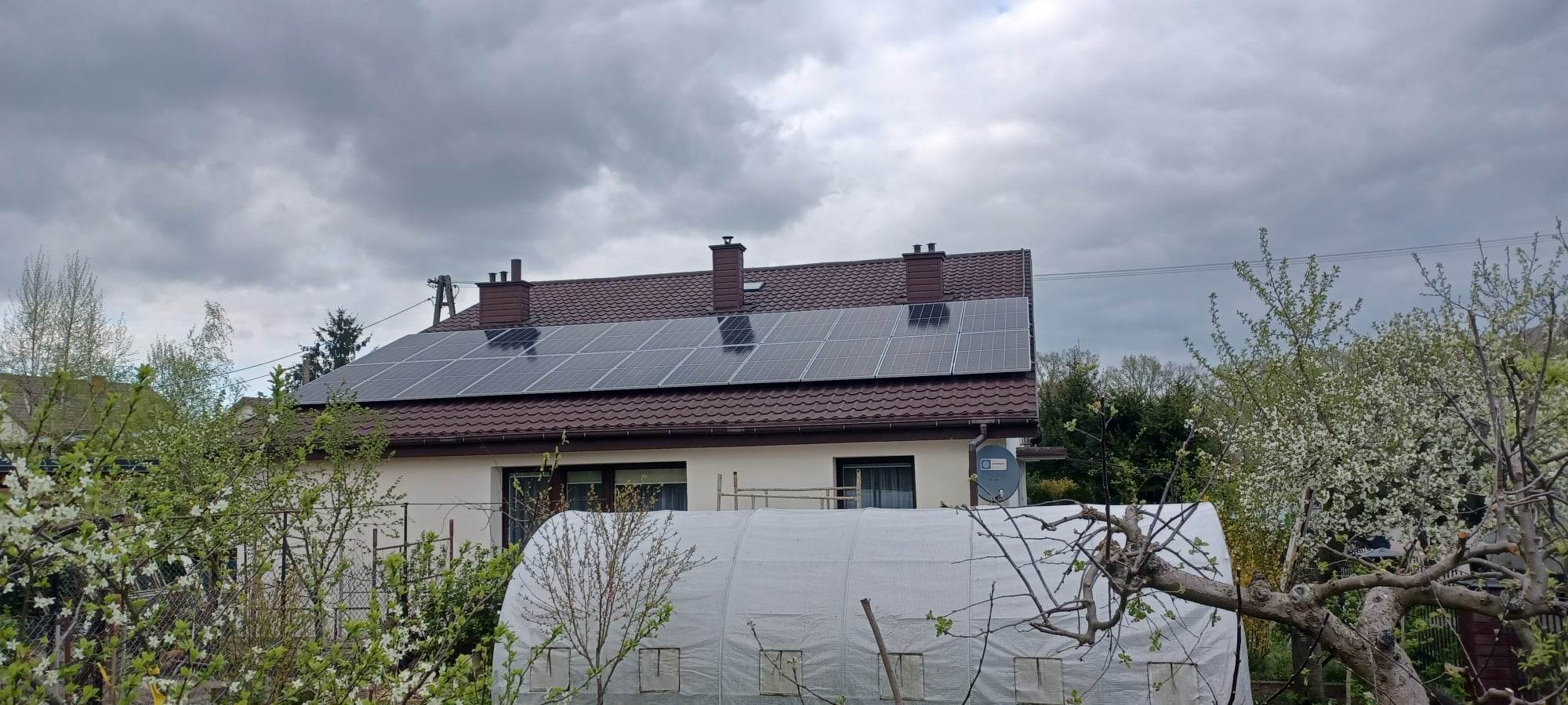 Panele słoneczne na brązowym dachu domu jednorodzinnego, widok z poziomu ogrodu z kwitnącymi drzewami owocowymi i szklarnią w tle, pochmurne niebo.