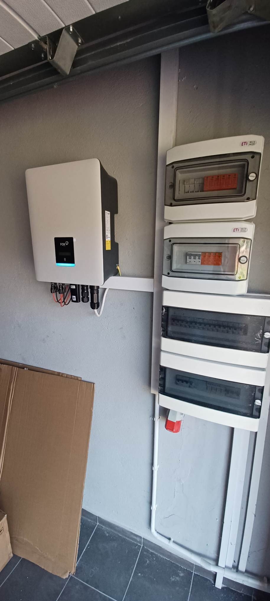 Zamontowany inwerter z podłączeniami kablowymi na szarej ścianie obok rozdzielnic elektrycznych ETI, widoczne kartony na podłodze.